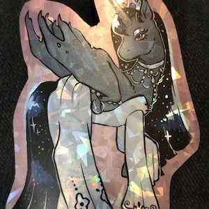 May include: A holographic sticker featuring a stylized illustration of a pony-like creature with a crown, horn, and flowing mane. The creature is primarily gray with a white dress and a starry cape. The sticker has a pink iridescent border.