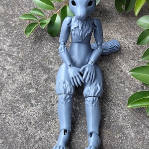May include: A grey articulated squirrel doll with a furry chest and tail, wearing ruffled trousers. The doll is seated on a stone surface, with green leaves in the background.