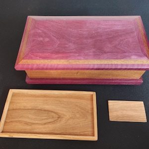 Puede incluir: Una caja de madera artesanal con una tapa morada vibrante y una base de madera natural. La caja es rectangular con una tapa con bisagras, y una bandeja separada y una pieza pequeña están al lado.