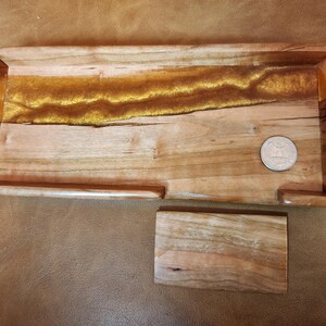 Puede incluir: Una bandeja rectangular de madera con un río de resina epoxi dorada que atraviesa el centro. Un pequeño posavasos de madera se encuentra debajo. Una moneda está en la bandeja como referencia de tamaño.