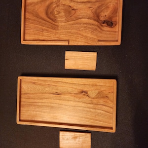 Puede incluir: Se muestran dos bandejas rectangulares de madera con bordes elevados y un pequeño bloque de madera. La madera tiene una veta natural y un color marrón claro y cálido. Una bandeja tiene un recorte en un lado. Los artículos son probablemente para servir o exhibir.