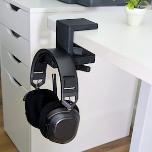 May include: A black headphone stand clamped to a white desk. The stand holds a pair of black over-ear headphones with soft earcups. The stand is made of a sturdy material and is designed to keep headphones organized and accessible.