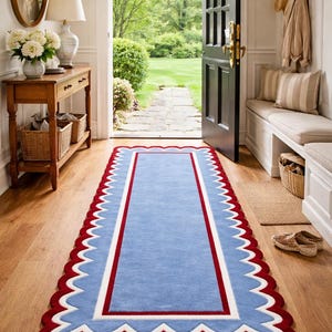 以下が含まれることがあります： 赤いスカラップの縁と白と赤の内側の縁が付いた、長方形の青いラグ。 ラグは、開いたドアから庭が見える、明るい玄関の木製の床に置かれています。