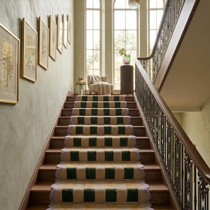 Puede incluir: Una gran escalera con escalones de madera y una alfombra con estampado verde y beige. Las paredes están pintadas de un verde suave y hay grabados botánicos enmarcados colgados en la pared. Una gran lámpara de araña cuelga del techo.