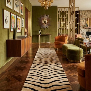 Op de afbeelding: Een interieur met een zebra-gestreept tapijt op een donkere houten vloer. De kamer heeft olijfgroene muren, kunstwerken en een gouden kroonluchter. Er zijn ook oranje en groene fluwelen stoelen. Het tapijt is ongeveer 300 cm lang.