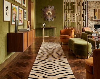 Puede incluir: Una alfombra de pasillo larga con un patrón de rayas de cebra en color canela y negro centrada sobre un suelo de madera oscura en espiga. La habitación tiene paredes verde oliva, un espejo sol dorado, una lámpara de araña y muebles de terciopelo.