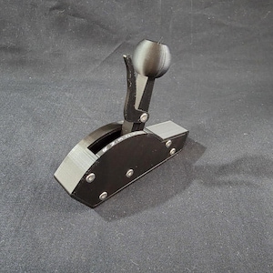 May include: A black 3D-printed automotive gear shifter assembly featuring a spherical knob, a vertical release lever, and a side plate with the printed letters R, N, 3, 2, 1. The mechanism is secured with visible silver-toned metal screws.