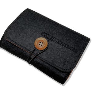May include: A black felt case for a Game Boy Advance, featuring a wooden button closure and the game console's name embossed on the front. The case is closed, with a rounded flap and a contrasting orange interior.