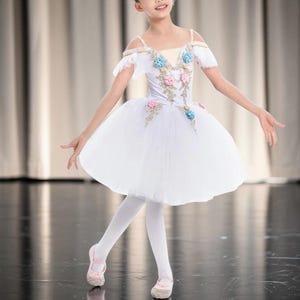 Może przedstawiać: Biała sukienka tutu do baletu z odkrytymi ramionami, ozdobiona różowymi i niebieskimi kwiatami. Sukienka ma warstwową tiulową spódnicę i jest połączona z białymi rajstopami i różowymi baletkami. Tancerka jest na scenie.