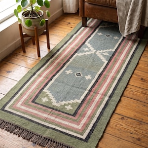 May include: A rectangular area rug featuring a muted green base with a central blue panel, accented by geometric patterns in white, pink, and black. The rug is finished with a fringed edge on the short side and displays a woven texture.