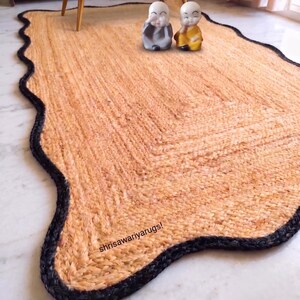 May include: A rectangular area rug with a natural tan woven texture and a thick, wavy black border. The rug features the text "shrisawariyarugs!" printed in small black letters near the bottom edge, providing a distinct contrast to the tan fibres.