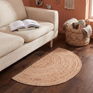 May include: A semi-circular area rug crafted from natural tan woven fibers. The rug features a concentric braided pattern and is positioned on a dark wood floor in front of a light beige sofa. The texture is coarse and organic in appearance.