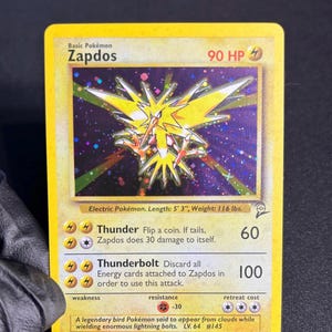 May include: A yellow Zapdos Pokémon card with a holographic background. The card features a yellow bird-like creature with lightning bolt wings. The card text includes "Thunder" and "Thunderbolt" attack descriptions, and the card is 5' 3" long and weighs 116 lbs.