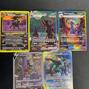 May include: Five Umbreon Pokémon trading cards. The cards feature Umbreon in various designs, including V, VMAX, and EX versions. The cards have different backgrounds and colours, with text and symbols indicating abilities and stats. The cards are in good condition.