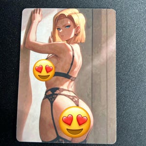 May include: A rectangular trading card featuring an illustration of a blonde character with blue eyes, wearing black lace lingerie with a garter belt. The character is positioned against a wall, looking over their shoulder. Two heart-eyed emojis cover parts of the image.