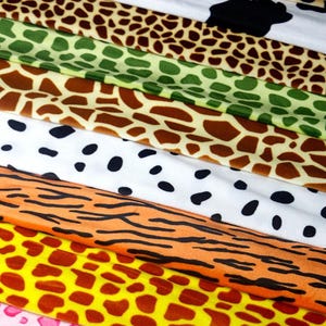 May include: A collection of fabric strips featuring various animal print patterns including giraffe, cow, dalmatian, tiger, and leopard designs in colors of brown, green, white, black, orange, yellow, and pink, stacked horizontally.