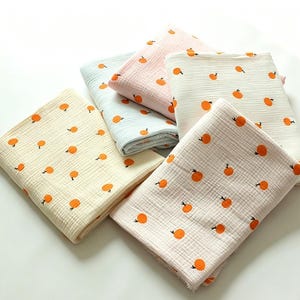 May include: Five folded muslin blankets in pastel shades of yellow, light blue, pink, white, and cream. Each blanket features a repeating pattern of small orange fruit icons with green stems scattered across the textured, lightweight fabric surface.
