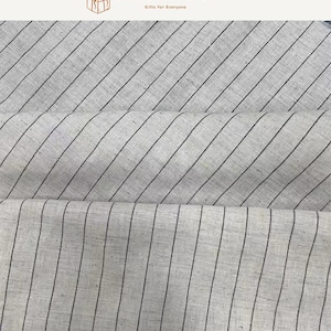 May include: A light beige fabric featuring a pattern of thin, dark brown vertical stripes. The material has a visible woven texture with natural variations in the weave. The fabric is folded into three horizontal layers, displaying the consistent striped design across the entire surface of the textile material.