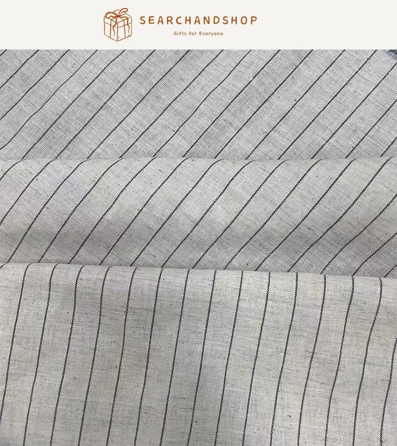 May include: A light beige fabric featuring a pattern of thin, dark brown vertical stripes. The material has a visible woven texture with natural variations in the weave. The fabric is folded into three horizontal layers, displaying the consistent striped design across the entire surface of the textile material.
