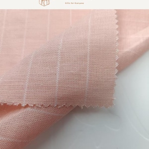 May include: A light pink fabric featuring a subtle white grid pattern. The textile has a visible woven texture with a soft, matte finish and a folded corner showing the raw, pinked edge. The top of the image includes the text SEARCHANDSHOP.
