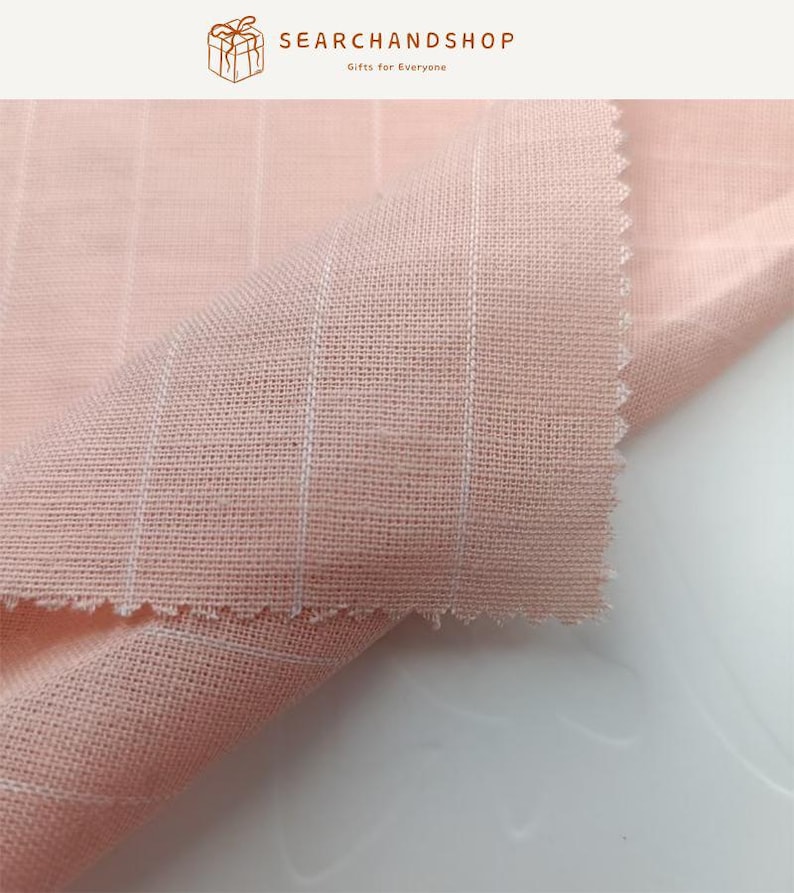 May include: A light pink fabric featuring a subtle white grid pattern. The textile has a visible woven texture with a soft, matte finish and a folded corner showing the raw, pinked edge. The top of the image includes the text SEARCHANDSHOP.