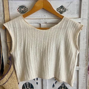 Op de afbeelding: Een mouwloze, gebroken witte top met een verticale ribbeltextuur en een brede, ronde halslijn. Het kledingstuk is afgewerkt met rafelige randen bij de armsgaten en de zoom en hangt aan een houten kledinghanger voor een wit uitgesneden houten paneel.