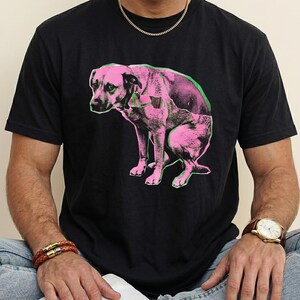 Może przedstawiać: Czarny t-shirt z różowo-zieloną grafiką przedstawiającą siedzącego psa. Grafika psa jest stylizowana w stylu pop-art. Koszulka ma okrągły dekolt i jest wykonana z miękkiego materiału.