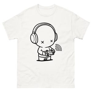 Puede incluir: Camiseta blanca con un gráfico de dibujos animados en blanco y negro. El gráfico representa un personaje con auriculares y sosteniendo un mando de videojuegos. El personaje tiene ojos en forma de 'x'. Una señal inalámbrica está al lado del personaje.