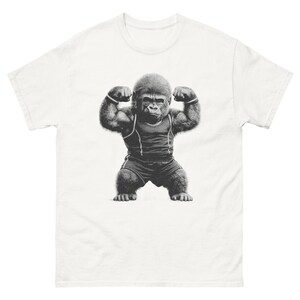 Puede incluir: Camiseta blanca con una ilustración en blanco y negro de un gorila musculoso flexionando sus bíceps. El gorila lleva una camiseta sin mangas y pantalones cortos oscuros, con muñequeras. El diseño está centrado en la parte delantera de la camiseta.