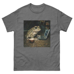 May include: Dark gray t-shirt featuring a graphic of a frog using a laptop with a coffee cup beside it. The frog is light brown and green with large eyes. The laptop is black and the coffee cup is brown.