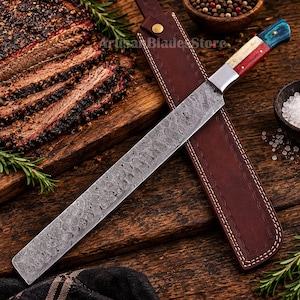 May include: A long, straight-edged knife with a patterned Damascus steel blade and a multi-colored handle featuring segments of white, red, and blue. The knife is accompanied by a brown leather sheath with a brass snap button closure.