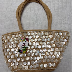 Puede incluir: Bolso tote de color beige con una textura tejida, adornado con numerosas lentejuelas redondas e iridiscentes. El bolso tiene asas beige a juego y una etiqueta con detalles florales y de texto. Las lentejuelas están dispuestas en el borde superior.