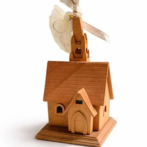 Puede incluir: Maqueta de iglesia de madera con una cruz en la parte superior. El modelo tiene un pequeño campanario y una cruz. Una cinta decorativa y tela están unidas a la cruz. La iglesia es de madera marrón claro.