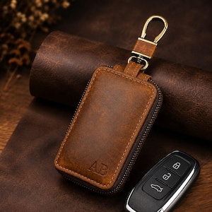 May include: A brown leather key case with a rectangular shape and a zipper closure. The front features the embossed letters AB. It includes a gold-toned metal swivel hook and a matching leather strap. A black car key fob is positioned nearby.