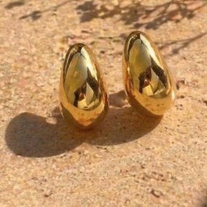 May include: A pair of gold-colored teardrop-shaped earrings. The earrings have a smooth, reflective surface, catching the light and reflecting the surroundings. The earrings are displayed on a textured, light-brown surface.