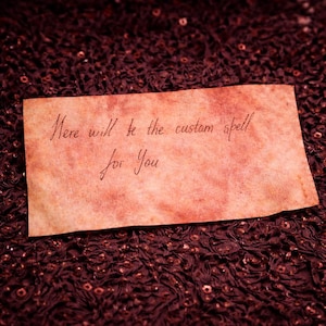 May include: A rectangular piece of aged paper with handwritten text in brown ink. The text reads "Here will be the custom spell for You". The paper has a reddish-brown hue and is set against a dark, textured background.