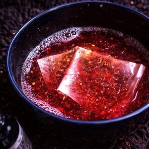 May include: A dark blue bowl filled with a reddish liquid and two tea bags. The liquid has bubbles and small particles. A dark bottle is in the lower left corner. The background is a dark, patterned fabric.