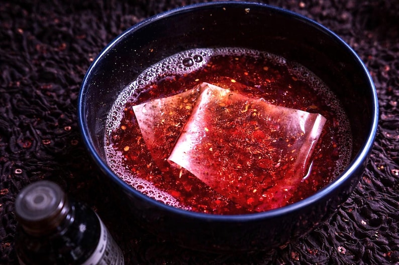 May include: A dark blue bowl filled with a reddish liquid and two tea bags. The liquid has bubbles and small particles. A dark bottle is in the lower left corner. The background is a dark, patterned fabric.