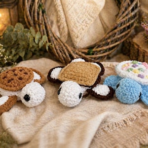May include: Three crocheted turtle toys. One is designed as a cookie sandwich, another as a toasted sandwich, and the third is blue with a colorful shell. Each turtle has white flippers and black eyes. The toys are on a beige surface.
