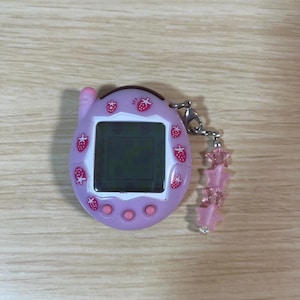 May include: A lavender handheld digital pet game with a black screen, adorned with red strawberry graphics. It has a pink antenna and three pink buttons. A silver chain is attached, dangling pink star-shaped beads.