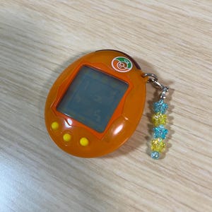 May include: Orange handheld digital pet game with a small screen and three yellow buttons. A silver chain is attached to the top with a charm of blue and yellow flower beads. The screen displays numbers.