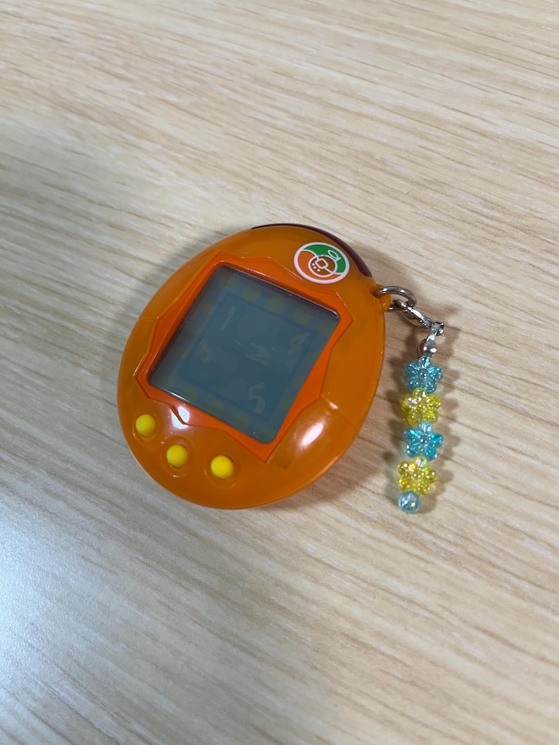 May include: Orange handheld digital pet game with a small screen and three yellow buttons. A silver chain is attached to the top with a charm of blue and yellow flower beads. The screen displays numbers.