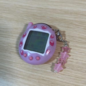 May include: A lavender handheld digital pet game with a screen displaying a pixelated graphic. The device is decorated with strawberry icons and has pink buttons. A silver chain with pink star charms is attached.