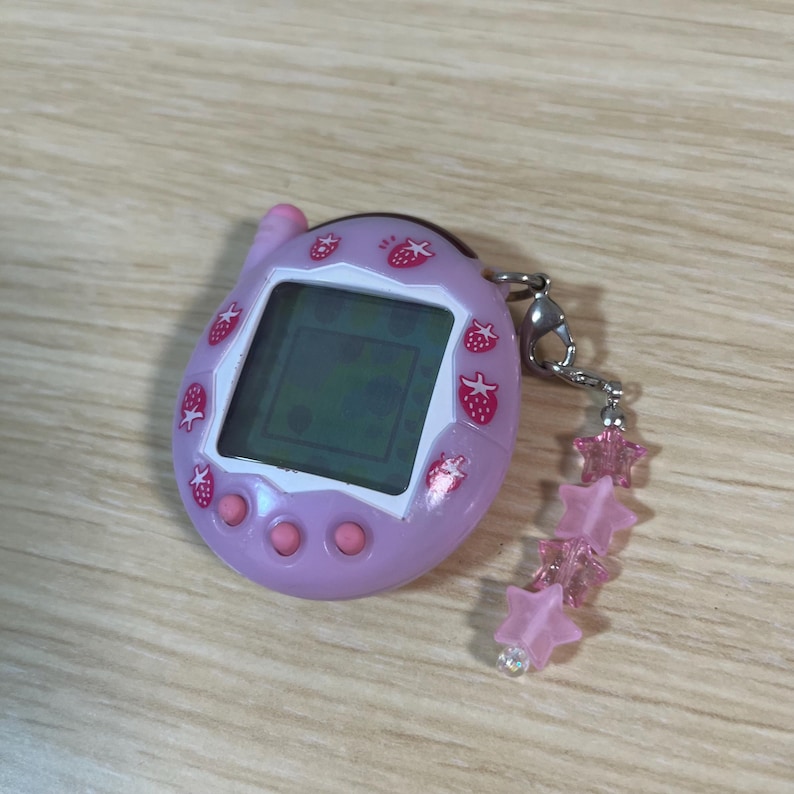 May include: A lavender handheld digital pet game with a screen displaying a pixelated graphic. The device is decorated with strawberry icons and has pink buttons. A silver chain with pink star charms is attached.