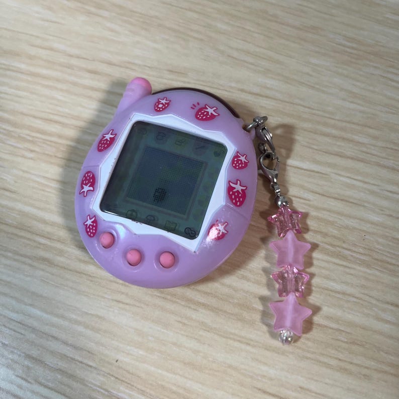 May include: A light purple handheld digital pet device with a screen and pink buttons. The device is decorated with strawberry designs. A charm with pink star-shaped beads hangs from the device.