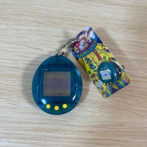 以下が含まれることがあります： 銀色のチェーンと3つの黄色のボタンが付いた、ティール色のTamagotchiバーチャルペットのおもちゃ。画面が表示され、新品未開封のTamagotchiがカラフルな段ボールの裏面に付いています。