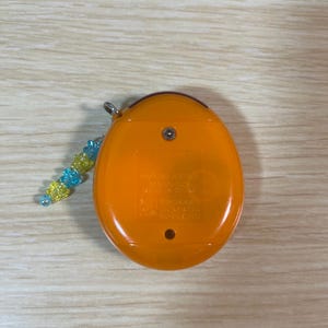 May include: An orange handheld electronic game with a small beaded charm. The device has a rounded shape with a small metal loop for the charm. The charm is made of blue and yellow beads.