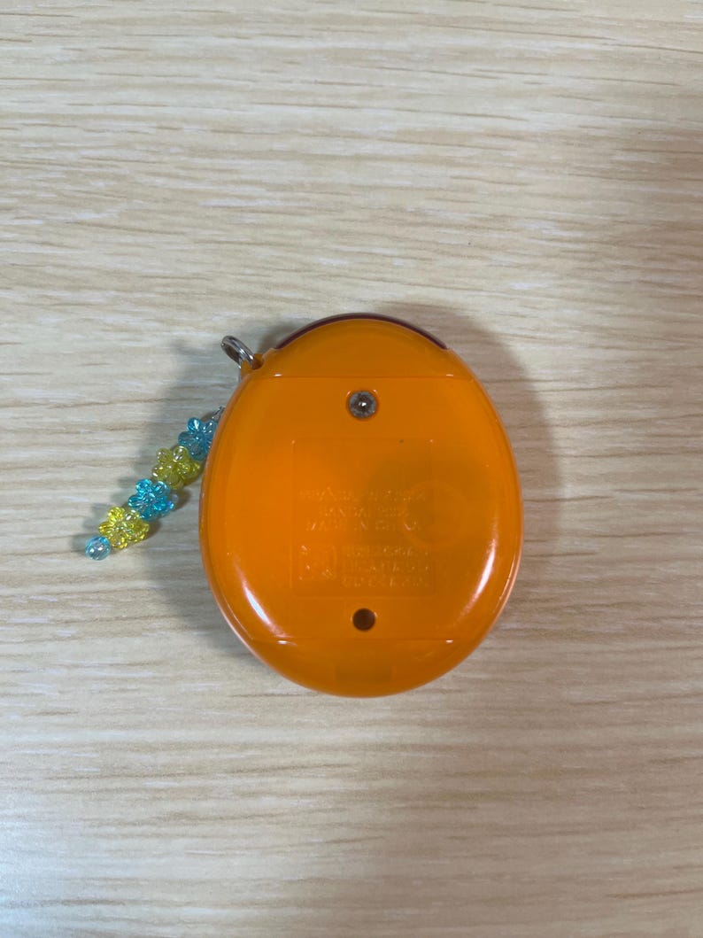 May include: An orange handheld electronic game with a small beaded charm. The device has a rounded shape with a small metal loop for the charm. The charm is made of blue and yellow beads.