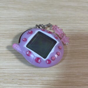 May include: A light purple handheld electronic game with a black screen and pink buttons. The device is decorated with pink strawberry graphics. A pink star-shaped charm hangs from a silver chain attached to the top.