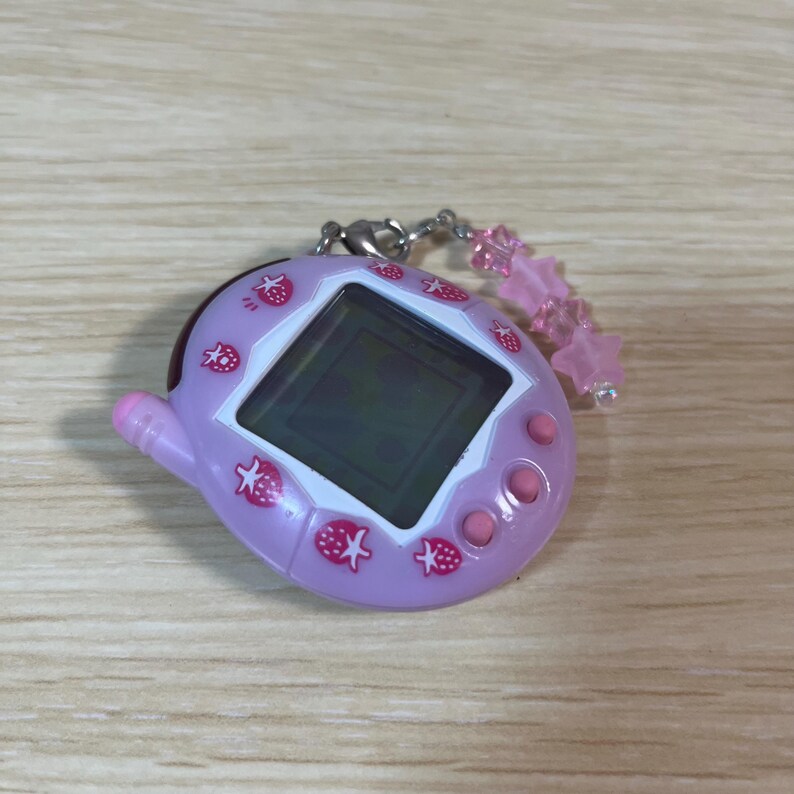 May include: A light purple handheld electronic game with a black screen and pink buttons. The device is decorated with pink strawberry graphics. A pink star-shaped charm hangs from a silver chain attached to the top.